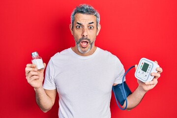 Handsome middle age man with grey hair using blood pressure monitor holding salt afraid and shocked...