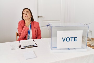 Beautiful middle age hispanic woman at political election sitting by ballot touching painful neck, sore throat for flu, clod and infection