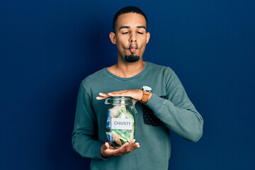 Young african american man holding charity jar with south africa rand banknotes making fish face with mouth and squinting eyes, crazy and comical.
