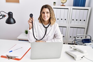 Young hispanic woman wearing doctor uniform holding stethoscope at medical clinic looking positive and happy standing and smiling with a confident smile showing teeth
