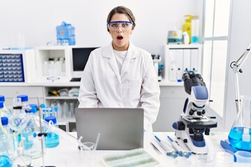 Young hispanic woman wearing scientist uniform working at laboratory scared and amazed with open mouth for surprise, disbelief face