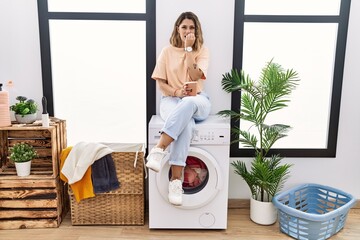 Young hispanic woman drinking coffee waiting for washing machine at laundry room looking stressed and nervous with hands on mouth biting nails. anxiety problem.