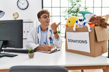 Young caucasian doctor man working at the clinic with toys donations box for children smiling happy...