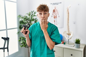 Young caucasian physio man holding hand grip to train muscle at the clinic covering mouth with hand, shocked and afraid for mistake. surprised expression