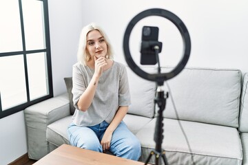 Fototapeta na wymiar Young caucasian woman recording vlog tutorial with smartphone at home looking confident at the camera with smile with crossed arms and hand raised on chin. thinking positive.