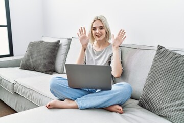 Young caucasian woman using laptop at home sitting on the sofa showing and pointing up with fingers number ten while smiling confident and happy.