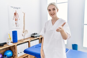 Young caucasian woman working at pain recovery clinic smiling cheerful offering palm hand giving assistance and acceptance.