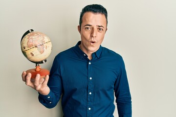 Handsome young man holding small world ball scared and amazed with open mouth for surprise,...