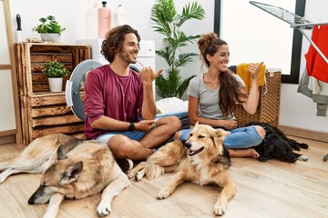 Young hispanic couple doing laundry with dogs smiling with happy face looking and pointing to the side with thumb up.