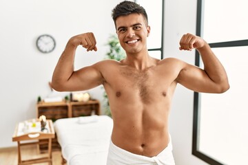Obraz premium Young hispanic man standing shirtless at spa center showing arms muscles smiling proud. fitness concept.
