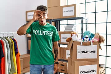 Young handsome hispanic man wearing volunteer t shirt at donations stand doing ok gesture shocked...