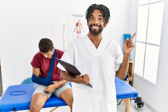 Young hispanic man working at pain recovery clinic with a man with broken arm with a big smile on face, pointing with hand finger to the side looking at the camera.