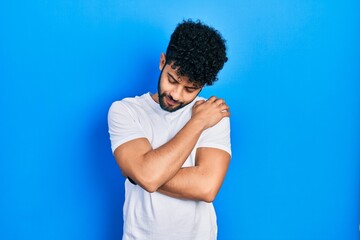 Young arab man with beard wearing casual white t shirt hugging oneself happy and positive, smiling...