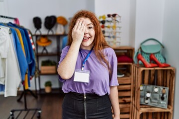 Young redhead woman working as manager at retail boutique covering one eye with hand, confident smile on face and surprise emotion.