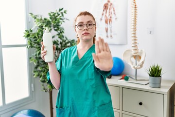 Young redhead physiotherapist woman holding massage body lotion with open hand doing stop sign with serious and confident expression, defense gesture