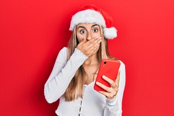 Beautiful hispanic woman wearing christmas hat holding smartphone covering mouth with hand, shocked...
