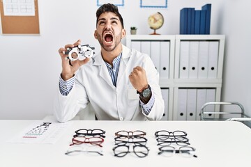Young optician man holding optometry glasses angry and mad screaming frustrated and furious,...