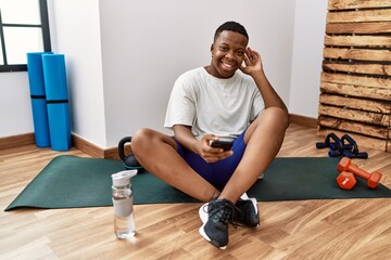 Obraz premium Young african man sitting on training mat at the gym using smartphone smiling with hand over ear listening an hearing to rumor or gossip. deafness concept.