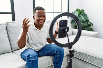Fototapeta na wymiar Young african man recording vlog tutorial with smartphone at home celebrating victory with happy smile and winner expression with raised hands