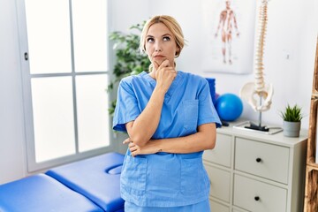 Beautiful blonde physiotherapist woman working at pain recovery clinic looking confident at the camera with smile with crossed arms and hand raised on chin. thinking positive.