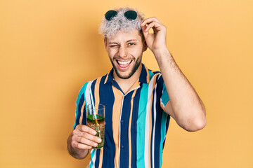 Young hispanic man with modern dyed hair drinking mojito glass winking looking at the camera with sexy expression, cheerful and happy face.