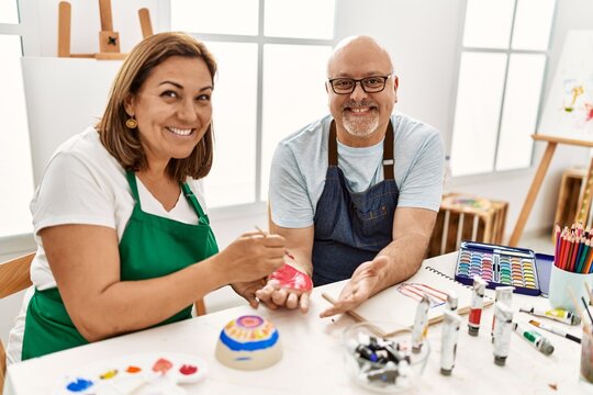 Middle age hispanic painter couple smiling happy painting palm hands at art studio.