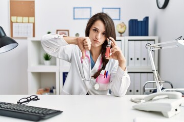Young doctor woman holding electronic cigarette at medical clinic with angry face, negative sign...