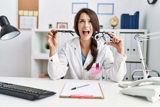 Young doctor woman holding optometry glasses and normal glasses angry and mad screaming frustrated and furious, shouting with anger looking up.