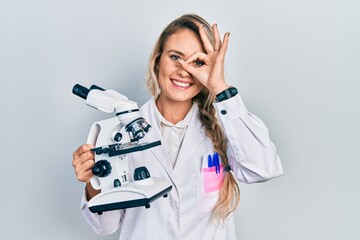 Beautiful young blonde woman holding microscope smiling happy doing ok sign with hand on eye...