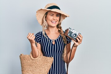 Beautiful young blonde woman holding vintage camera winking looking at the camera with sexy...