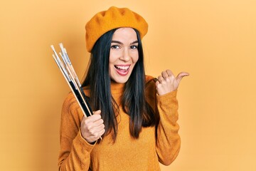 Young brunette woman wearing artist look with beret holding paintbrushes pointing thumb up to the...