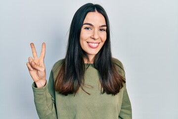 Young brunette woman wearing casual green sweater smiling with happy face winking at the camera doing victory sign. number two.