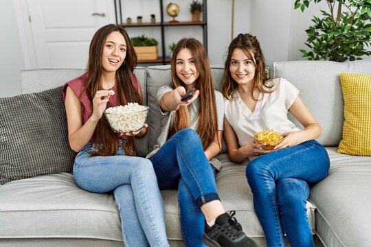 Three young hispanic woman smiling happy watching film eating popcorn and chips potatoes at home. - Powered by Adobe