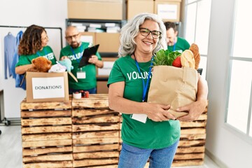 Group of middle age volunteers working at charity center. Woman smiling happy and holding paper bag with food to donate.
