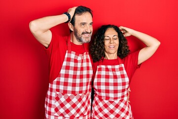 Middle age couple of hispanic woman and man wearing cook apron smiling confident touching hair with hand up gesture, posing attractive and fashionable