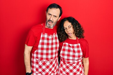 Middle age couple of hispanic woman and man wearing cook apron in shock face, looking skeptical and sarcastic, surprised with open mouth