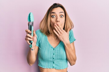 Young caucasian blonde woman holding silicone spatula covering mouth with hand, shocked and afraid...