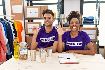 Young interracial people wearing volunteer t shirt at donations stand showing and pointing up with fingers number six while smiling confident and happy.