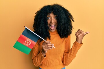 African american woman with afro hair holding afghanistan flag pointing thumb up to the side smiling happy with open mouth