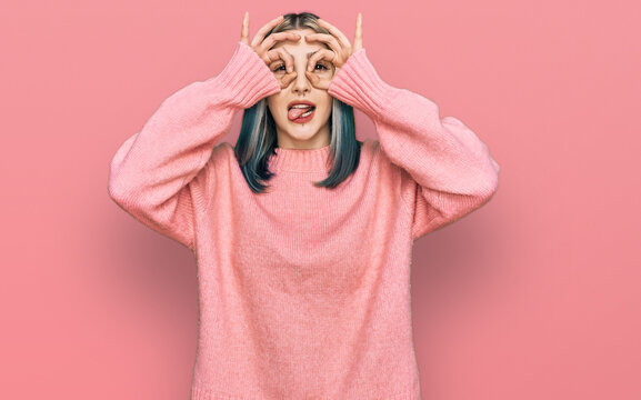 Young modern girl wearing pink wool winter sweater doing ok gesture like binoculars sticking tongue out, eyes looking through fingers. crazy expression.