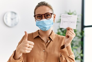 Young blonde woman holding covid record card smiling happy and positive, thumb up doing excellent...