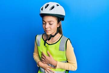 Beautiful brunette little girl wearing bike helmet and reflective vest with hand on stomach because indigestion, painful illness feeling unwell. ache concept.