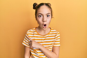 Beautiful brunette little girl wearing casual striped t shirt surprised pointing with finger to the side, open mouth amazed expression.