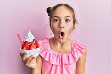 Beautiful brunette little girl eating strawberry ice cream scared and amazed with open mouth for...
