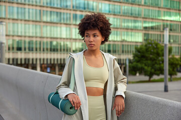 Pensive young woman leads sporty lifestyle carries rolled fitness mat concentrated away with thoughtful expression dressed in sportswear poses against city background. Sport and recreation concept