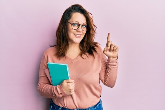 Young caucasian woman holding book smiling with an idea or question pointing finger with happy face, number one
