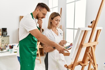 Fototapeta premium Young caucasian couple with serious expression drawing at art studio. Man standing and looking draw canvas.