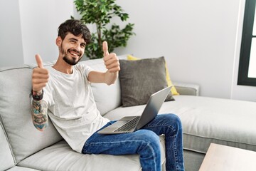 Hispanic man with beard sitting on the sofa approving doing positive gesture with hand, thumbs up smiling and happy for success. winner gesture.