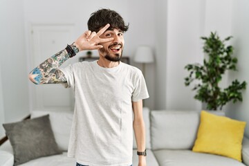 Hispanic man with beard at the living room at home doing peace symbol with fingers over face, smiling cheerful showing victory