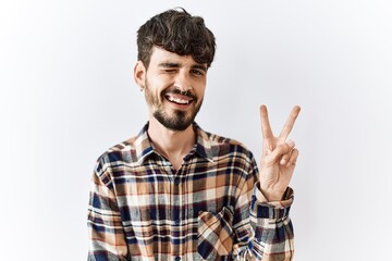 Fototapeta premium Hispanic man with beard standing over isolated background smiling with happy face winking at the camera doing victory sign. number two.